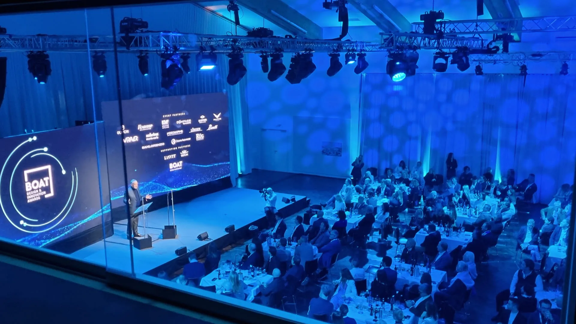 Ein Redner auf einer Konferenz vor Publikum in blau beleuchtetem Raum.